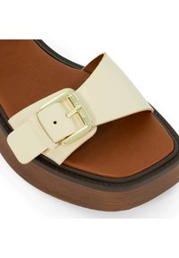 Crème-kleurige slipper met een brede band, metalen gesp-accent, en een bruine, gestructureerde voetbed op een chunky platformzool.