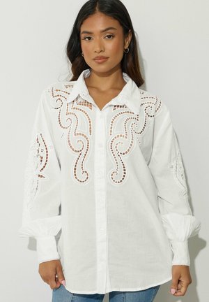 Kobieta ubrana w białą bluzkę z długim rękawem z misternymi ażurowymi wzorami z przodu i na rękawach, w połączeniu z niebieskimi dżinsami.