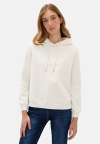 Giovane donna che indossa un felpa con cappuccio beige chiaro con lacci e jeans blu, in posa su uno sfondo semplice.