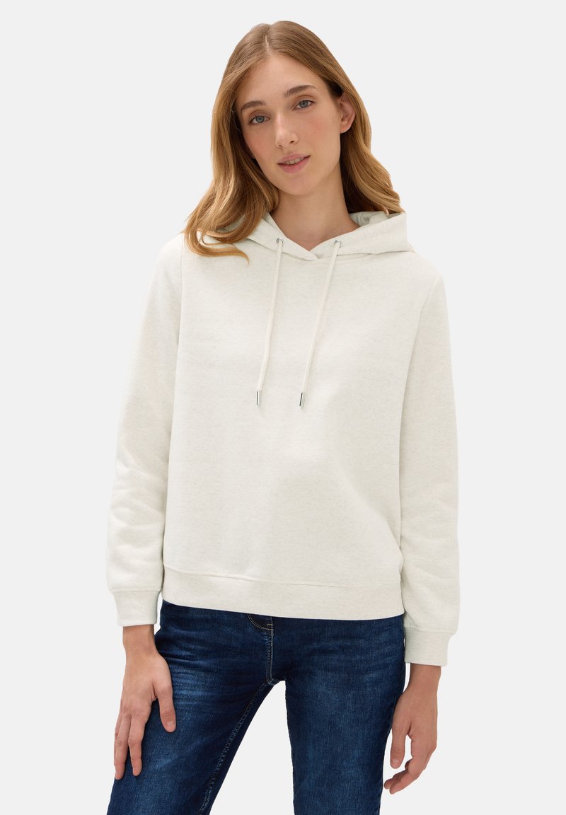 Giovane donna che indossa un felpa con cappuccio beige chiaro con lacci e jeans blu, in posa su uno sfondo semplice.