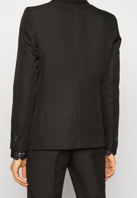 Blazer nero su misura con colletto strutturato, maniche lunghe e due bottoni ai polsini. Il tessuto ha una texture liscia e un design aderente.