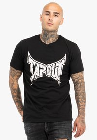 Melnā kokvilnas T-krekls ar lielu, nodilušu balto "TAPOUT" logo priekšpusē. Īsas piedurknes un apaļais kakls.