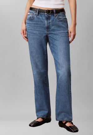 Femme portant un jean bleu droit, ceinture noire, haut blanc et chaussures plates noires, debout devant un fond gris.