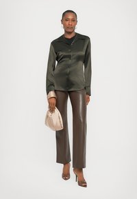 Camicia verde oliva in satin con bottoni, pantaloni in ecopelle marroni e una morbida borsa beige. Il modello indossa tacchi alti e orecchini semplici.