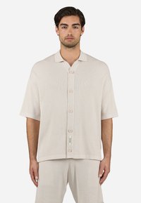 Camisa de manga corta de botonadura en tela de canalé beige claro, con cuello, cinco botones en la parte delantera y una etiqueta en el dobladillo.