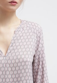 Blouse gris clair avec un décolleté en V, présentant un motif géométrique en blanc et rose doux. Le tissu a une texture lisse et des manches longues.