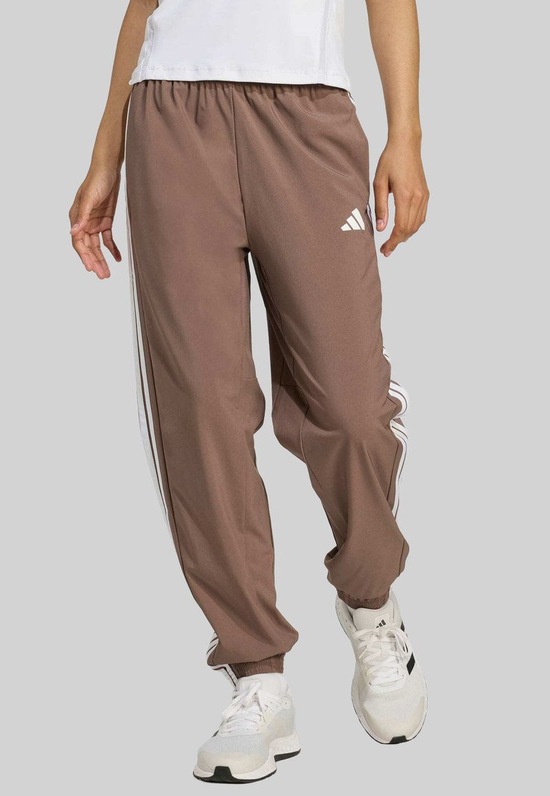 Brown polyester track pants with white side stripes, elastic waistband, and ankle cuffs. Paired with white athletic shoes.