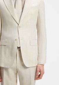 Homme portant un costume crème à rayures fines, à deux boutons, poches à rabat, une cravate assortie et un mouchoir blanc dans la poche.