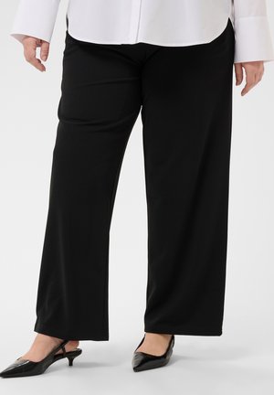Persona che indossa pantaloni neri a gamba larga, una camicia bianca e scarpe slingback nere con tacco basso, in piedi su una superficie bianca.