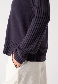 Personne portant un pull côtelé violet foncé avec les mains dans les poches d’un pantalon côtelé blanc sur un fond uni.