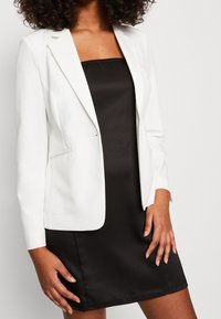 Blazer blanc ajusté à manches longues, revers crantés et poches avant, porté sur une robe nuisette en satin noir. Les textures lisses des tissus sont visibles.