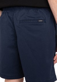Marinblå shorts tillverkade av lättviktsmaterial, med resår i midjan och en bakficka med en liten svart etikett.