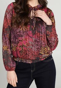 Blusa a maniche lunghe, morbida, realizzata in tessuto trasparente con motivi floreali nei colori rosa, viola e giallo, dotata di scollo a fiocco e orlo arricciato.