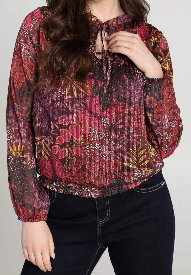 Blusa a maniche lunghe, morbida, realizzata in tessuto trasparente con motivi floreali nei colori rosa, viola e giallo, dotata di scollo a fiocco e orlo arricciato.