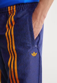 Mørkeblå treningsbukser med oransje striper, et geometrisk mønster, lett stoff, sidelomme og brodert logo på låret.