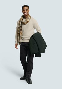 Beige trui, geruite sjaal in bruintinten, donkere broek en zwarte laarzen. Houdt een groene jas vast. Glimlachend, staande op een lichte achtergrond.