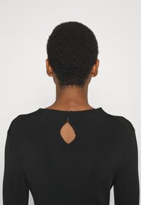 Personne aux cheveux courts et bouclés portant un haut noir avec une petite découpe en forme de goutte d'eau à l'arrière près de l'encolure.