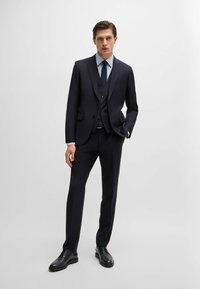 Abito blu navy a tre pezzi con revers a punte, gilet a tre bottoni e pantaloni slim-fit. Abbinato a una camicia a quadri blu chiaro e scarpe nere.