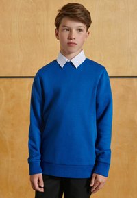 Felpa blu con maniche lunghe e collo tondo, con una camicia bianca a colletto sotto. Texture liscia, indossata sopra pantaloni neri.