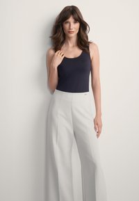 Kobieta z długimi brązowymi w łosami, ubrana w czarny top bez rękawów i jasnoszare spodnie z wysokim stanem i szerokimi nogawkami, stoi na tle białej ściany.