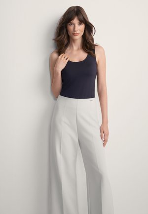 Kobieta z długimi brązowymi włosami, ubrana w czarny top bez rękawów i jasnoszare spodnie z wysokim stanem i szerokimi nogawkami, stoi na tle białej ściany.