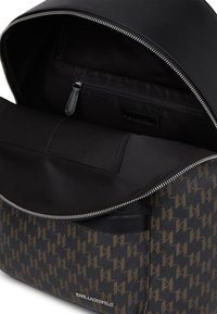 Fekete hátizsák struktúrált dizájnnal, barna logómintával. Cipzáras záródás, texturált anyagok és belső zsebből álló részletek.