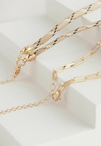 Drei Halsketten auf einer weißen Oberfläche präsentiert: ein gewebtes Design in Beige und Schwarz, eine goldene Kette mit Gliedern und polierter goldener Hardware.