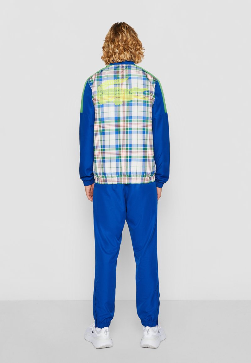 Tenue de sport colorée comprenant une veste bleue à motif écossais avec des poignets élastiques, associée à un pantalon bleu assorti et des baskets blanches.