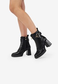 Stivaletti neri in pelle con chiusura a zip, tacco quadrato spesso e un accento decorativo in oro sulla parte superiore. Texture liscia, punta arrotondata.