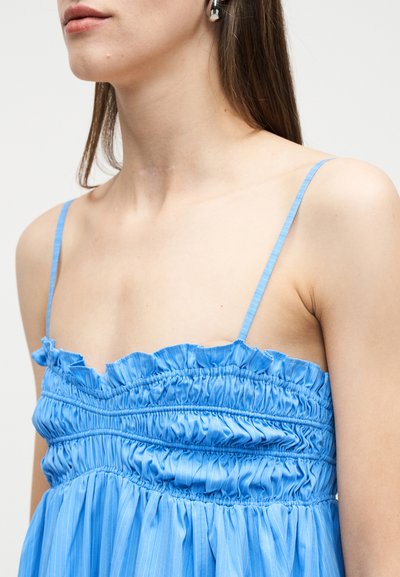 Κοντινό πλάνο γυναίκας που φοράει μπλε τοπ με τιράντες σπαγγέτι, ραφές ρυτιδώσεις, με βολάν στο ντεκολτέ και διακριτικές κάθετες ρίγες.