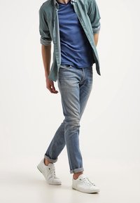 Ljusblå jeans med tapered fit, uppvikta fållar, kombinerat med en mörkblå t-shirt och en ljusgrön skjorta med krage, accentuerad av vita sneakers.