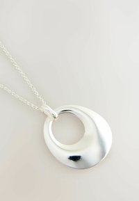 Silverpendant på en kedja, med en slät, rund design och ett öppet centrum. Den polerade ytan reflekterar ljuset, vilket förstärker dess form.