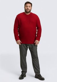 Rode gebreide trui met een gestructureerd patroon, ronde halslijn en lange mouwen, gecombineerd met grijze geruite broek en zwarte schoenen.