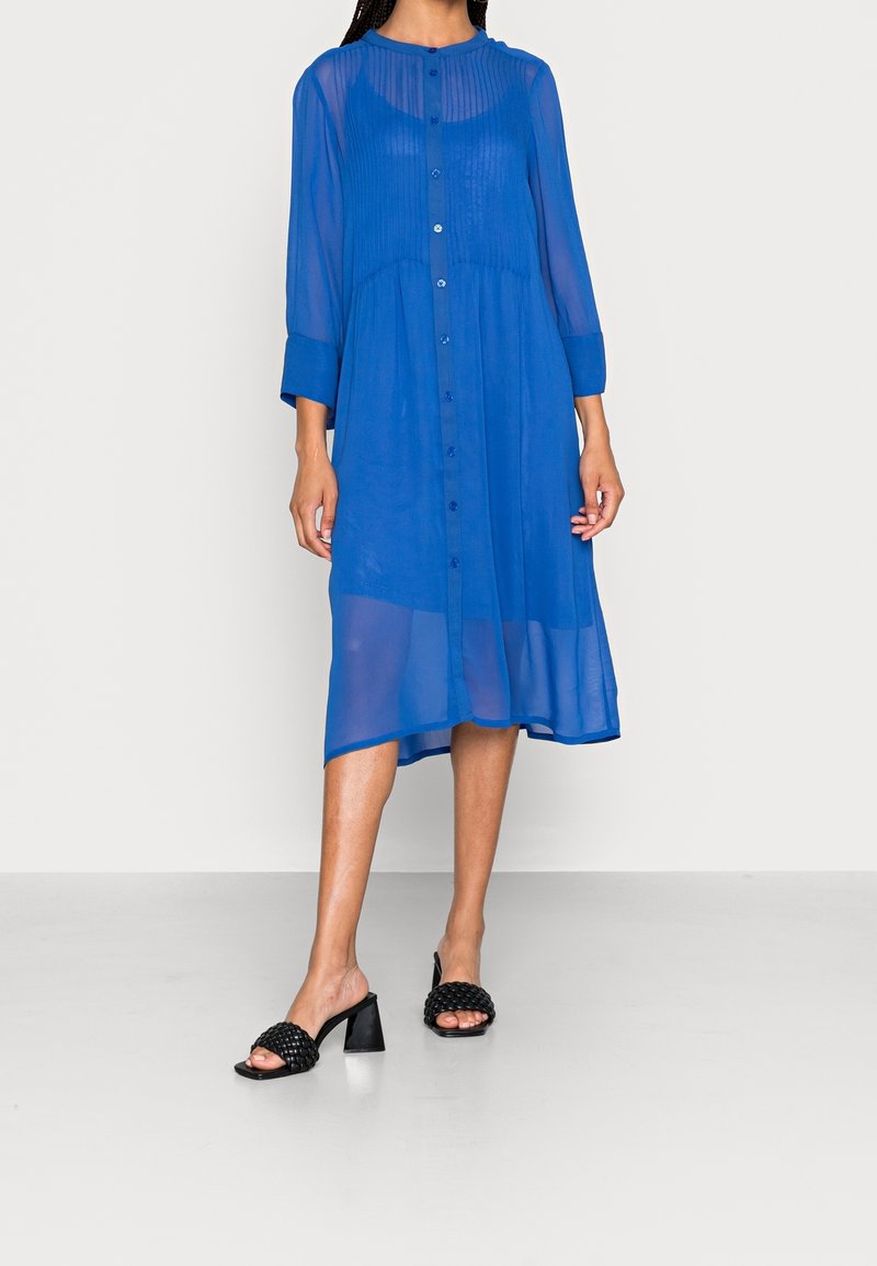 Femme portant une robe bleu roi transparente, boutonnée, longueur genou, avec manches longues et sandales à talons noires.