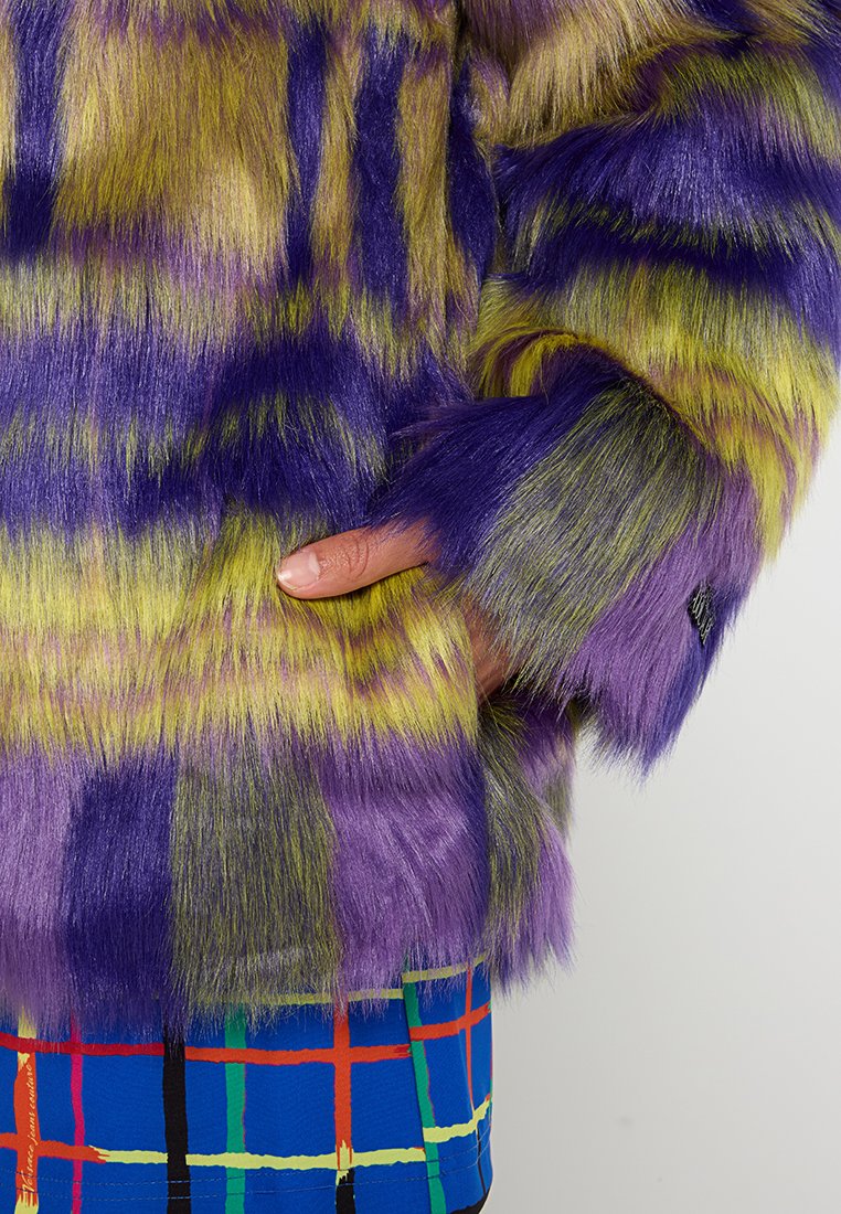Faux-Felljacke in lila und grünen Streifen, mit langem, weichem Material. Eine Hand greift am Handgelenk in das Fell und zeigt die lebendigen Farben.