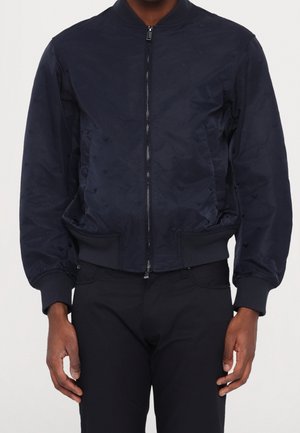 Personne portant une veste bomber bleu marine foncé unie zippée avec poignets et ourlet côtelés, et un pantalon foncé sur un fond uni.