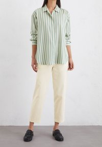 Camicia a maniche lunghe a righe verde chiaro e bianco, con colletto e maniche arrotolate. Abbinata a pantaloni color crema e mocassini neri.