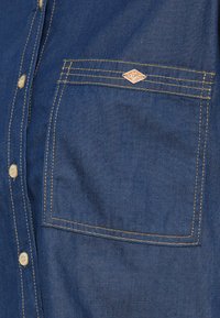 Σκούρο μπλε πουκάμισο από τζιν με τσέπη στο στήθος, έντονη κίτρινη ραφή και μικρό έμβλημα. Κουμπιά μπροστά με στρογγυλά κουμπιά.