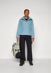 Svetlo modra vodoodporna jakna s kapuco, kombinirana s črnimi cargo hlačami in svetlo sivo nahrbtnikom s črnimi poudarki, nošena s strani stojeca modela.