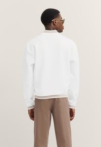 White jacket with ribbed cuffs and collar, featuring subtle tan stripes. Smooth texture, casual silhouette, paired with brown trousers.