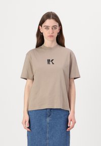 Bež majica s kratkimi rokavi in okroglim izrezom, s črnim tiskanim logotipom "K" na prsih, izdelana iz gladkega bombažnega materiala.