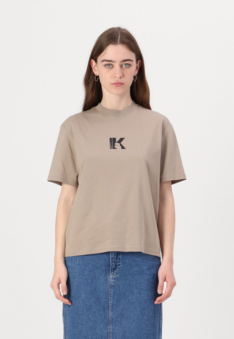 Bež majica s kratkimi rokavi in okroglim izrezom, s črnim tiskanim logotipom "K" na prsih, izdelana iz gladkega bombažnega materiala.