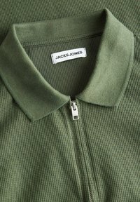 Vihreä polo-paita, jossa on tekstuuri, vetoketjullinen etuosa ja tasainen kaulus. Siinä on valkoinen etiketti, jossa on "JACK&JONES" -brändäys.