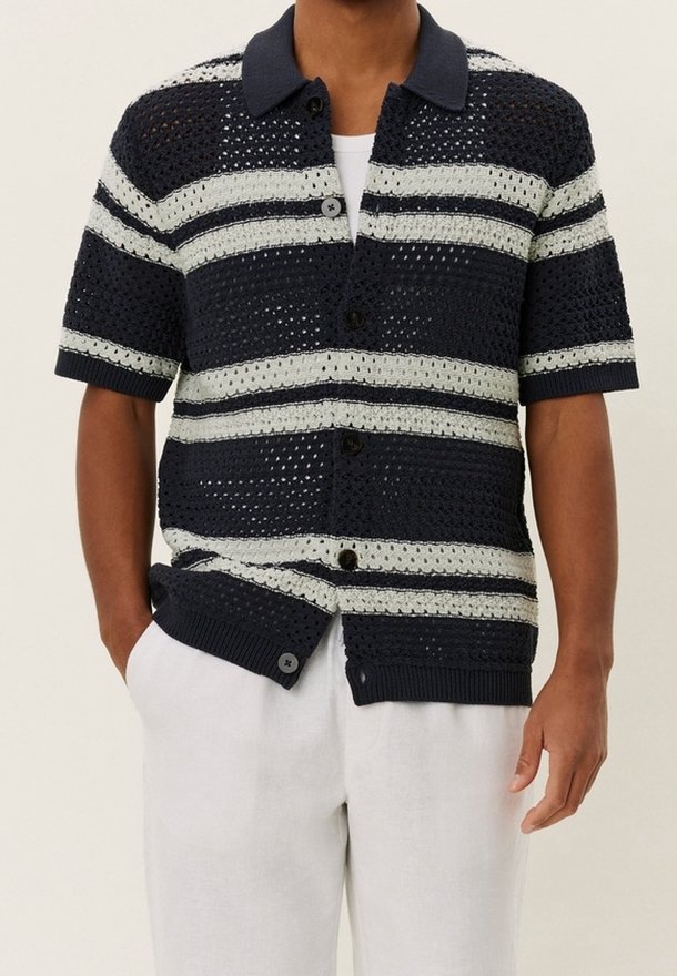 Homme portant un pull en maille à manches courtes rayé bleu marine et gris avec des boutons sur un sous-maillot blanc et un pantalon blanc, mains dans les poches.