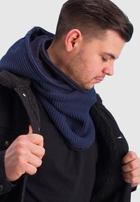 Navy knitted scarf with a textured pattern, layered under a black fleece-lined jacket. The scarf is draped around the neck.