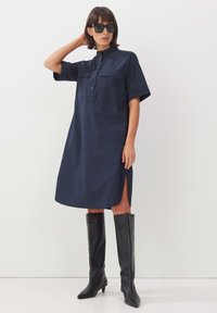 Navyblaues Hemdkleid mit Stehkragen, kurzen Ärmeln und Knopfleiste. Verfügt über zwei Brusttaschen und seitliche Schlitze. Kombiniert mit schwarzen kniehohen Stiefeln.