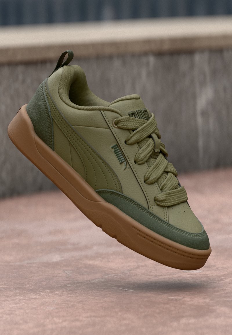 Zelena i tamnozelena Puma tenisica s smešnim đonom, vezice zavezane, lebdi iznad ružičaste podloge pored pozadine od teksturiranog kamenog zida.