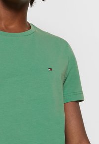 T-shirt en coton vert avec un col rond. Présente un petit logo aux couleurs rouge, blanc et noir sur la poitrine gauche. Manches courtes.