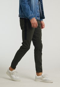 Olivgröna tapered byxor med svarta sidopaneler, gjorda av mjukt tyg. Matchas med vita sneakers. Man i en jeansjacka går.