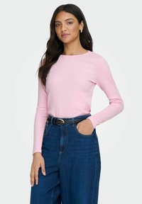 Roze langslavende shirt met een ronde hals, gecombineerd met hoge blauwe denim jeans en een zwarte ceintuur. Over het geheel een gladde textuur.